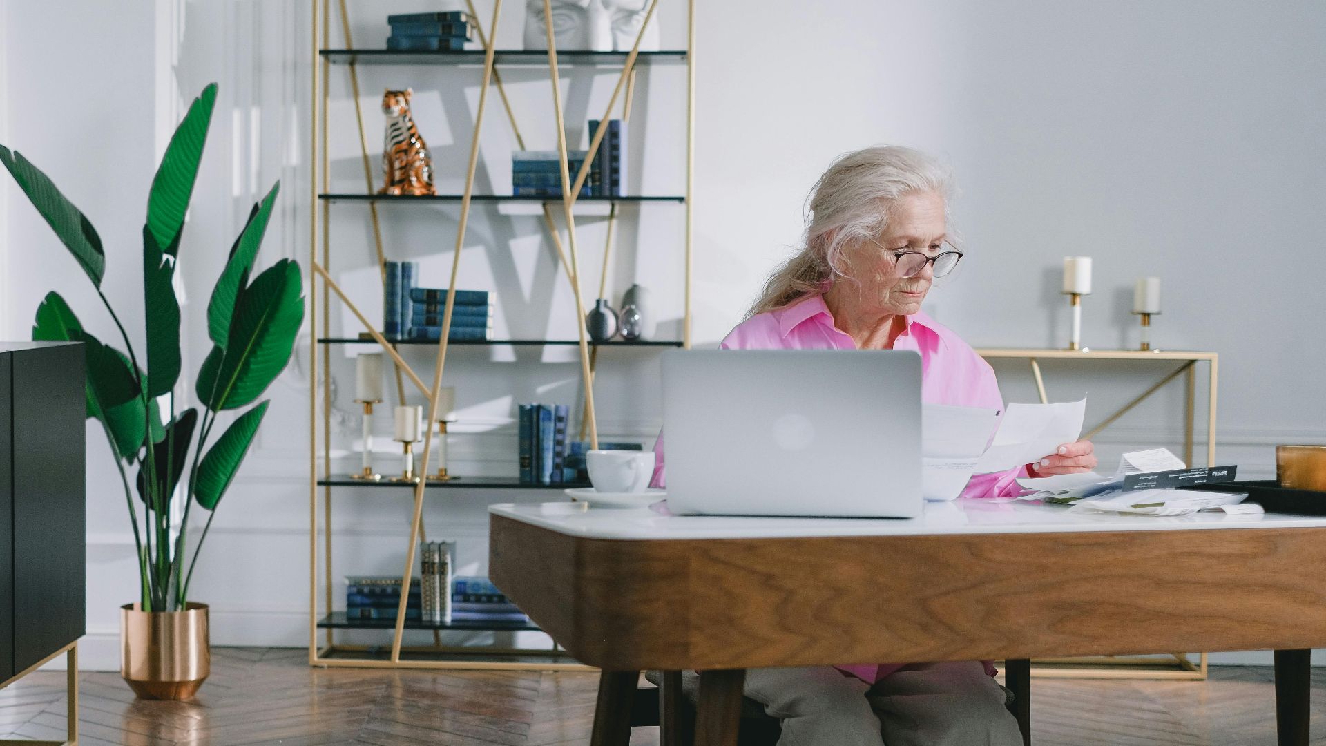Elderly woman reading papers at a desk with a laptop in a stylish home office setting.