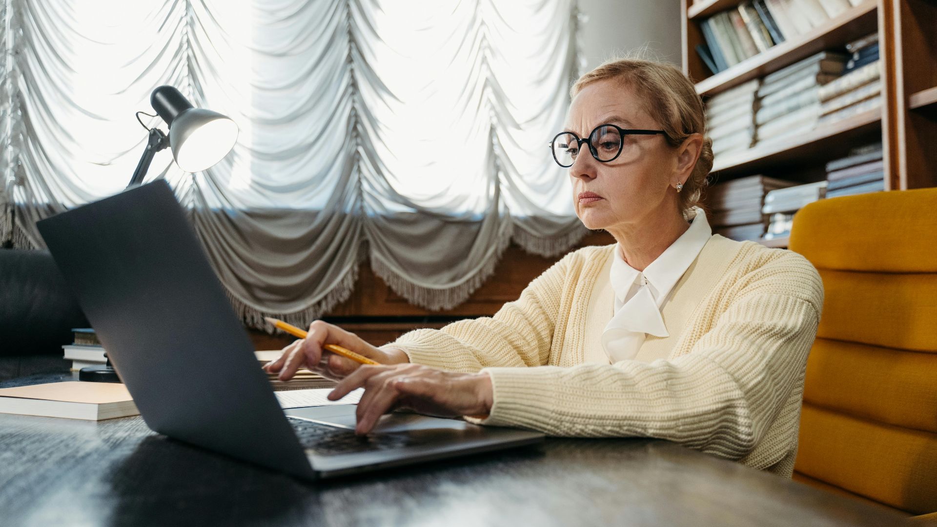 Mature woman in glasses working intently on a laptop at a library desk.