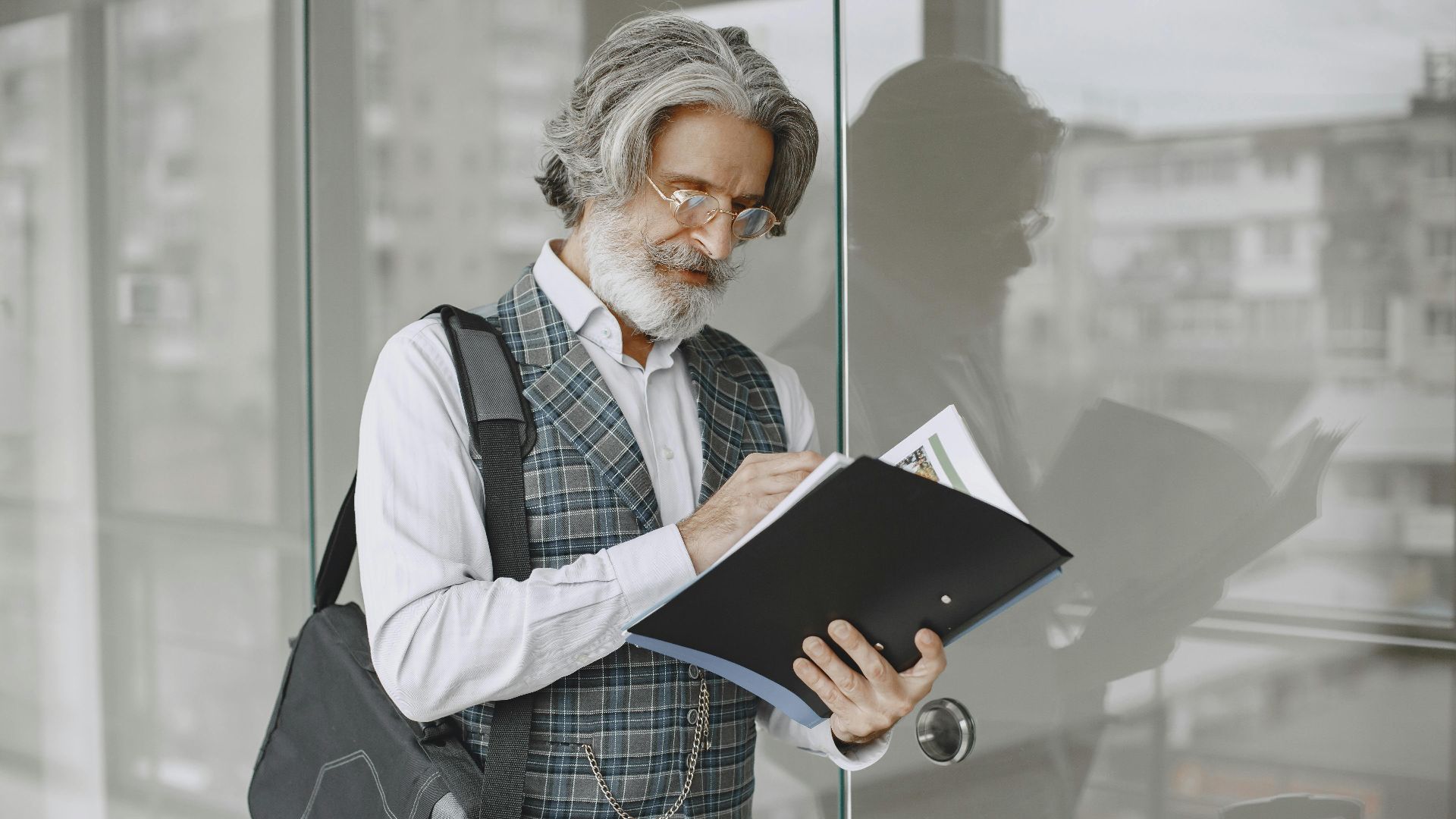 Elderly man in stylish office attire reading documents by glass windows.