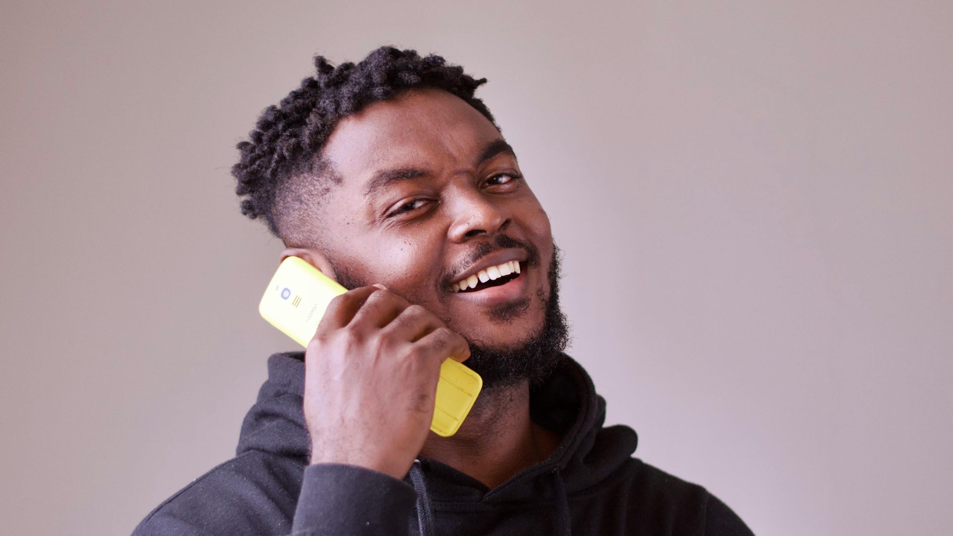 Happy man wearing a black hoodie, talking on a yellow mobile phone indoors, captured in a studio setting.