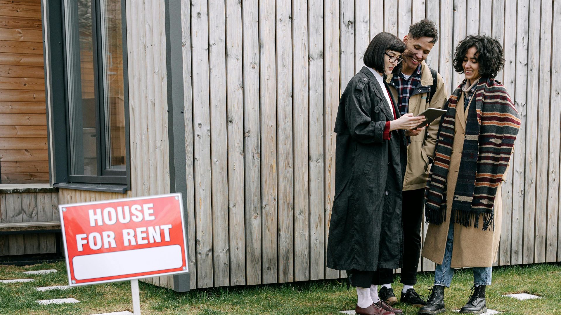 A group of three people reviewing a tablet near a 'House for Rent' sign outside.
