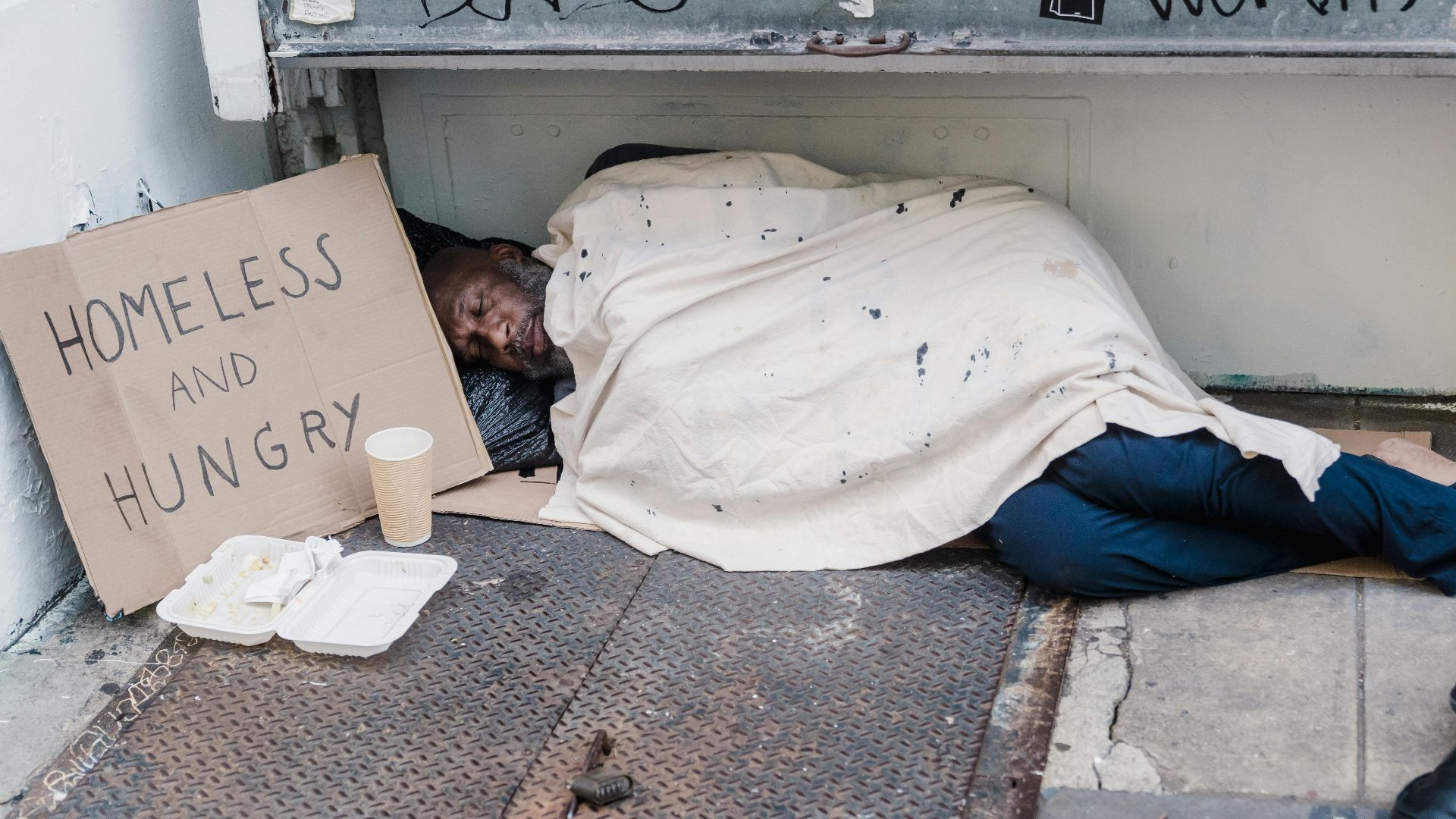 Street scene depicting a homeless man sleeping with a placard reading 'Homeless and Hungry'.