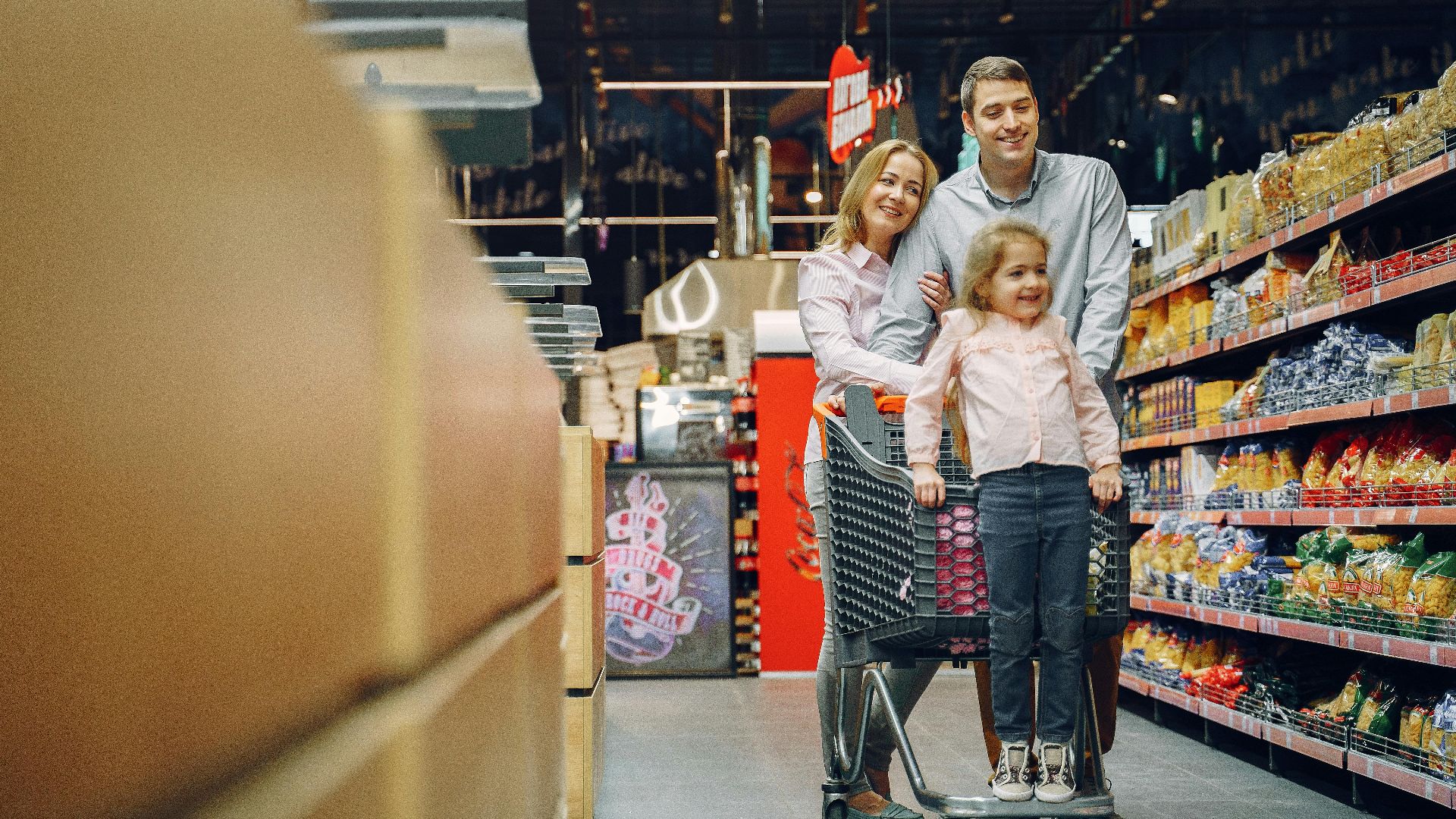 Family enjoying a shopping trip in a supermarket, with a child in a cart and parents smiling.