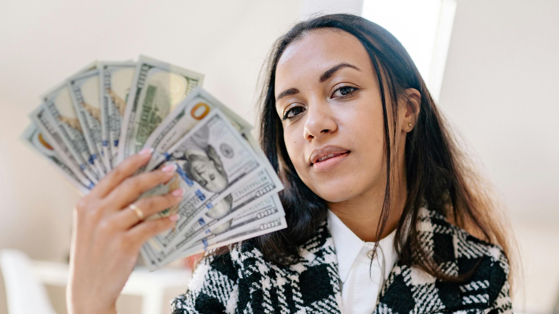 Woman holding American dollars confidently, depicting wealth and success in a bright indoor setting.