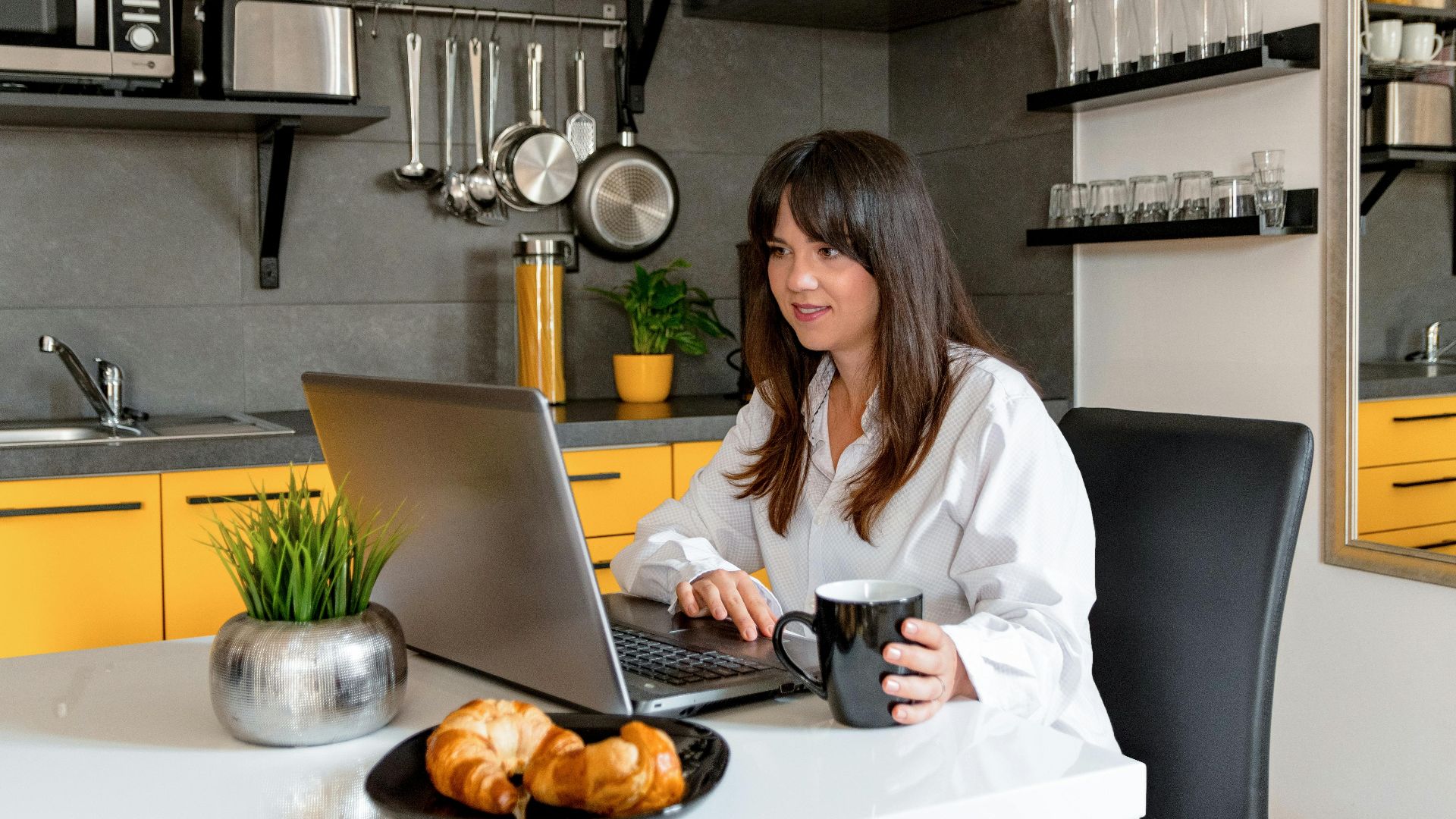 A woman in a kitchen wearing white long sleeves, holding a mug, and using a laptop.