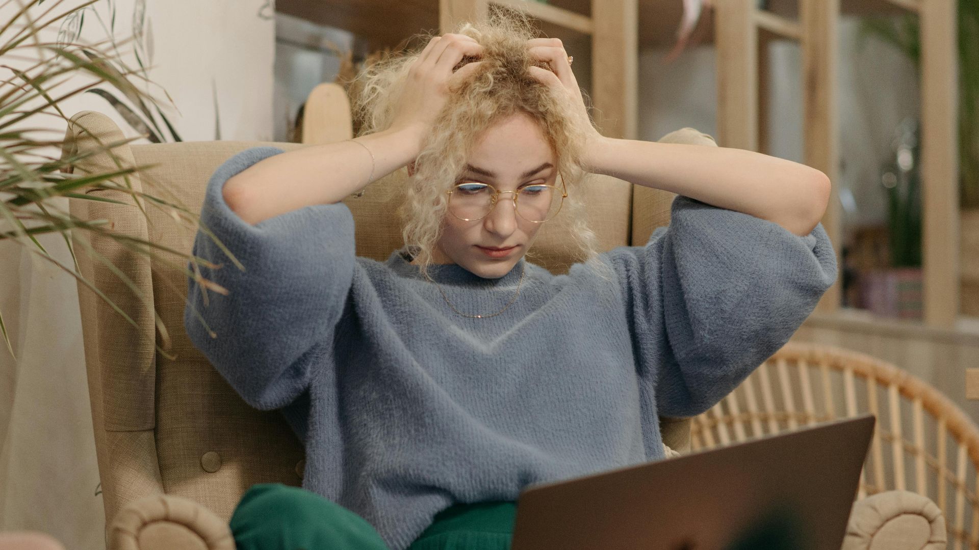 A young woman looking stressed while using her laptop indoors, sitting in an armchair with hands on her head.