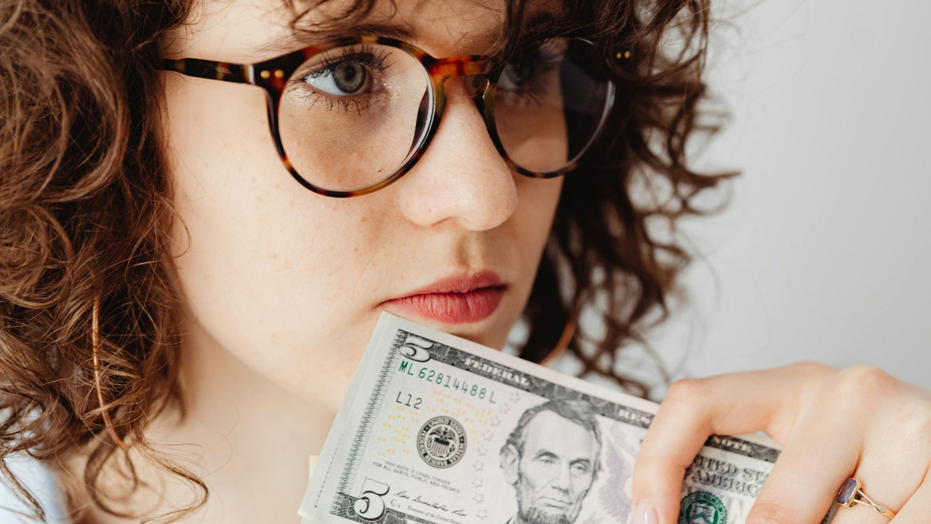 Close-up of a woman holding a US five dollar bill, wearing glasses and looking thoughtful.