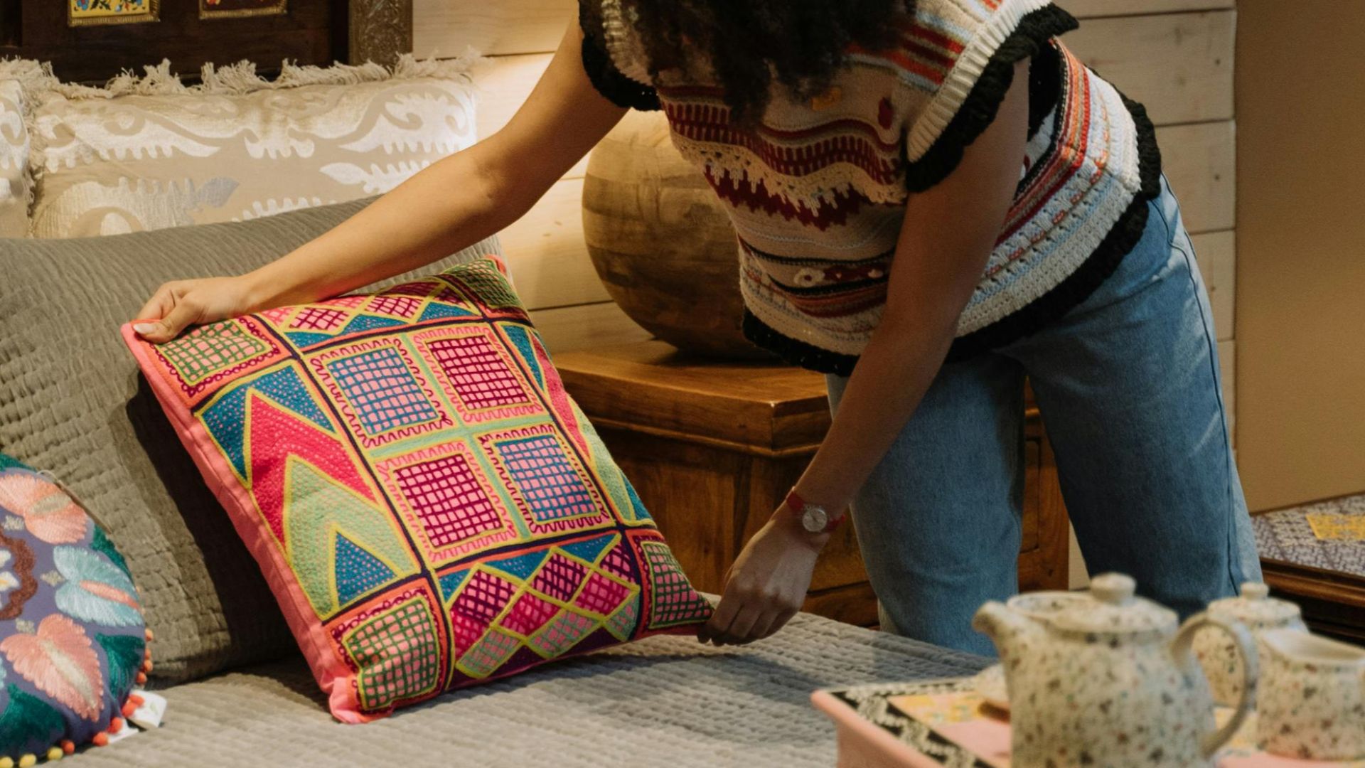 A woman arranges colorful pillows in a bohemian style bedroom featuring a tea set and decorative elements.