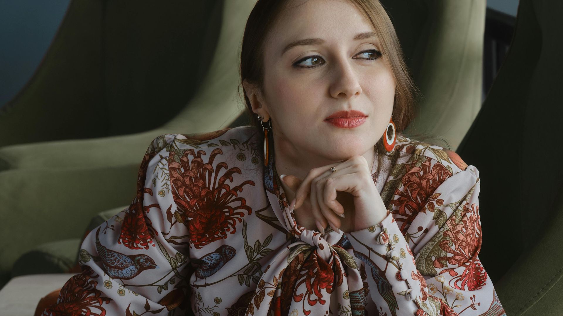 Contemplative woman in stylish floral blouse reading a magazine indoors, seated on a modern chair.