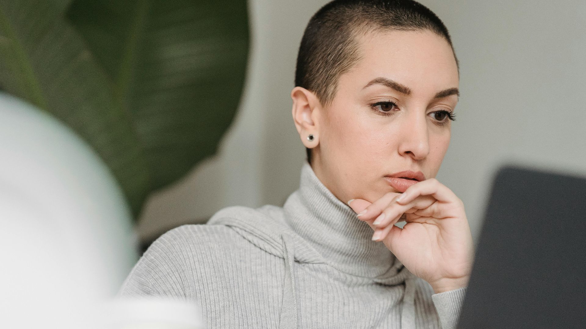 Thoughtful female with short hairstyle looking at netbook screen and touching chin in light room