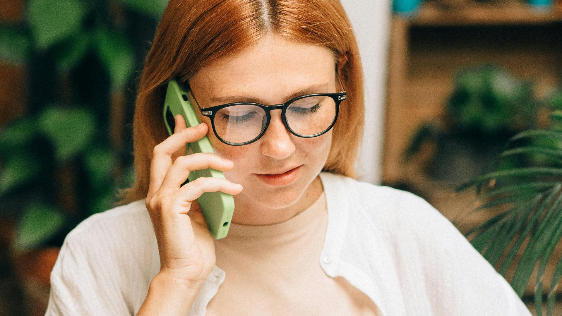 Woman in glasses discussing document on phone surrounded by plants indoors.
