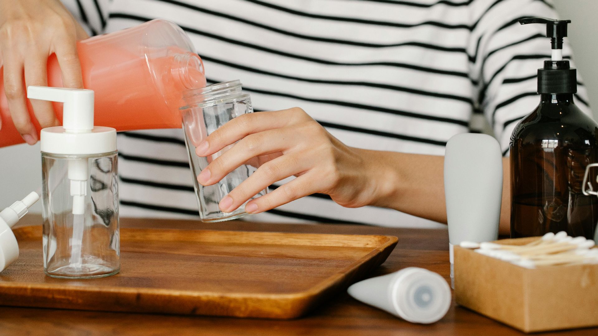 Crop person sitting at table and pouring peach coloured soap from opened soap dispenser into plastic bottle
