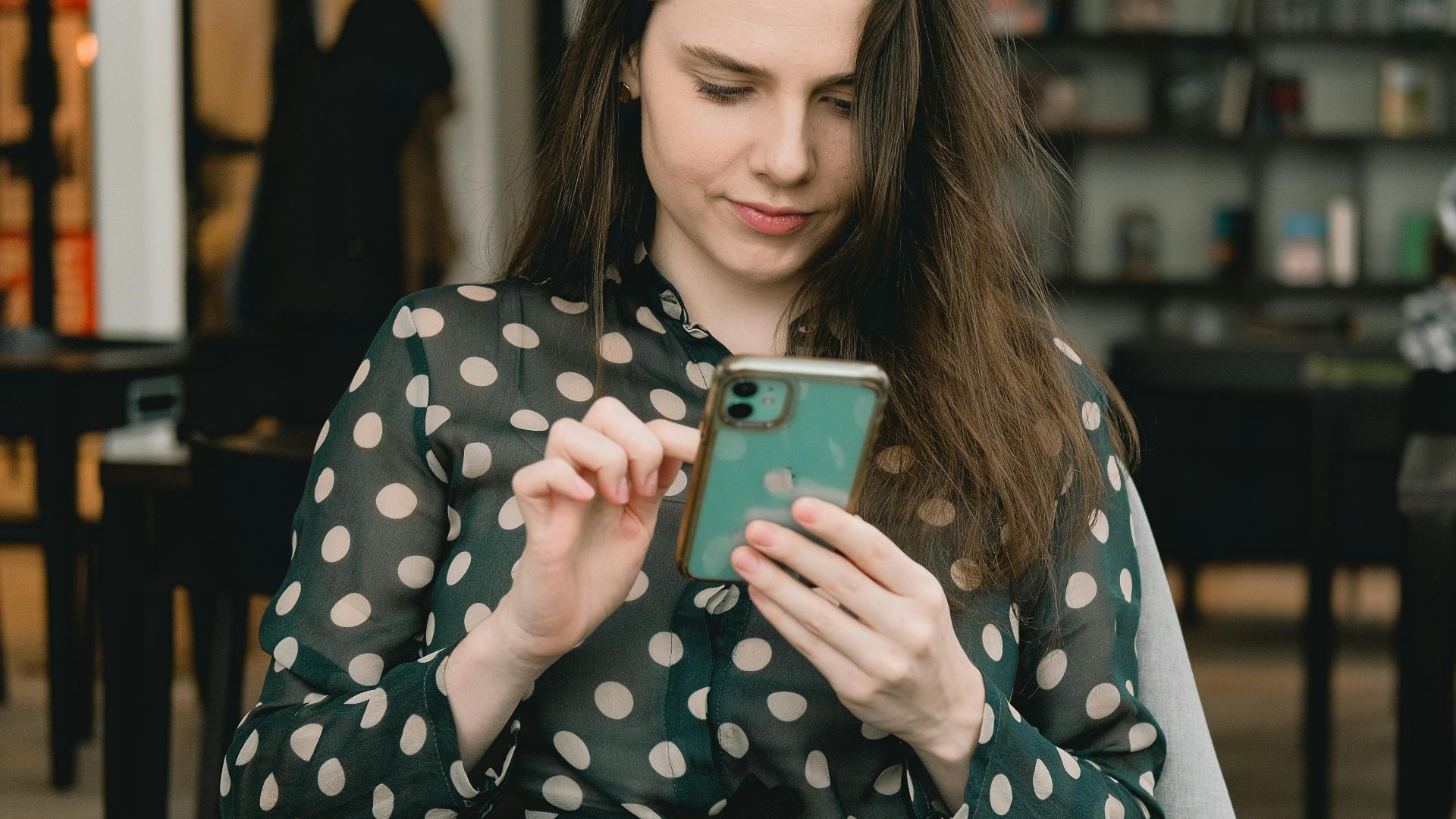 Woman in polka dot dress using smartphone indoors at a library. Cozy and modern setting.