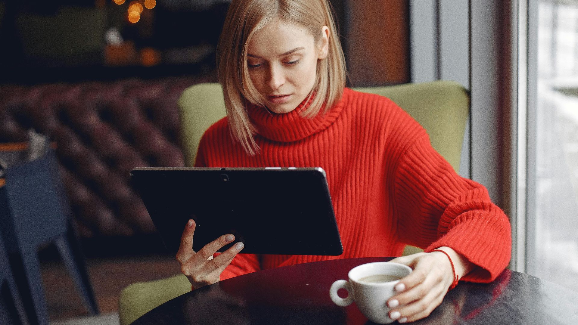 Woman in a red sweater sitting and using a tablet with a cup of coffee in a cozy café.