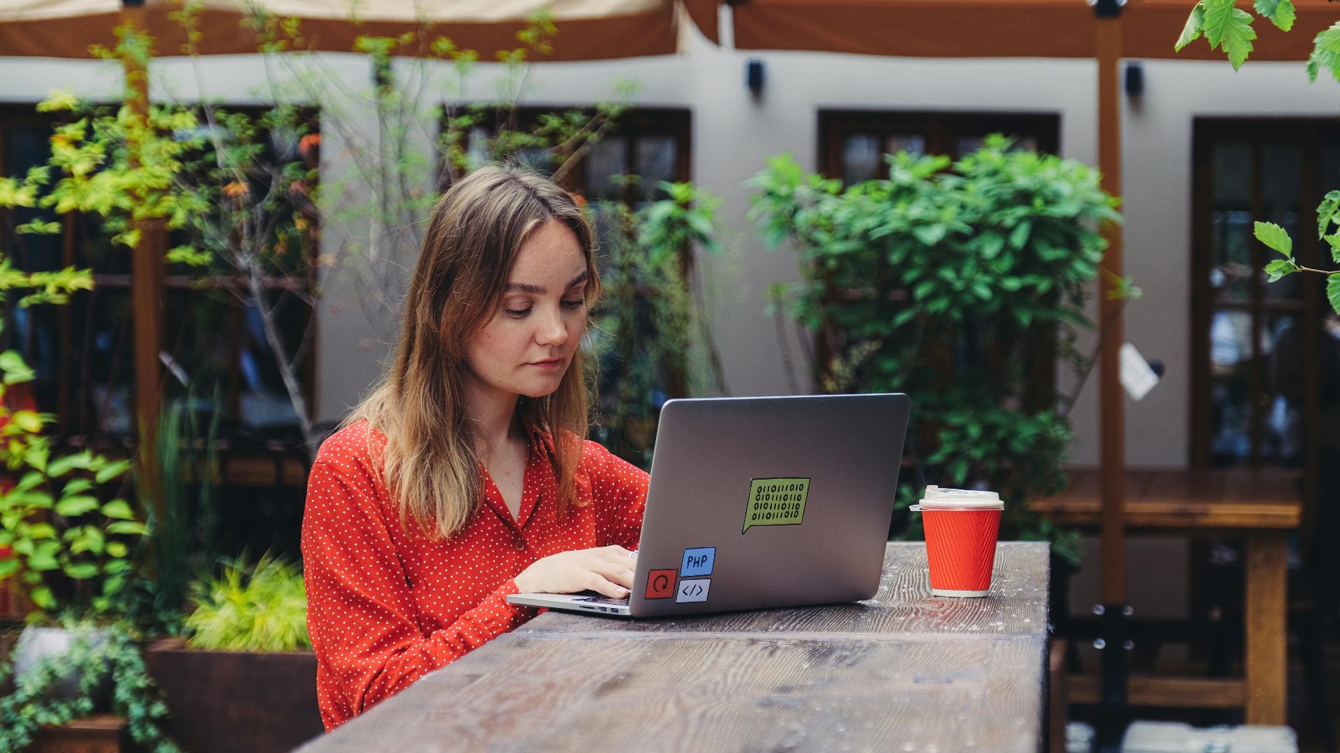 Young woman working on a laptop in an outdoor cafe setting, enjoying a cup of coffee.