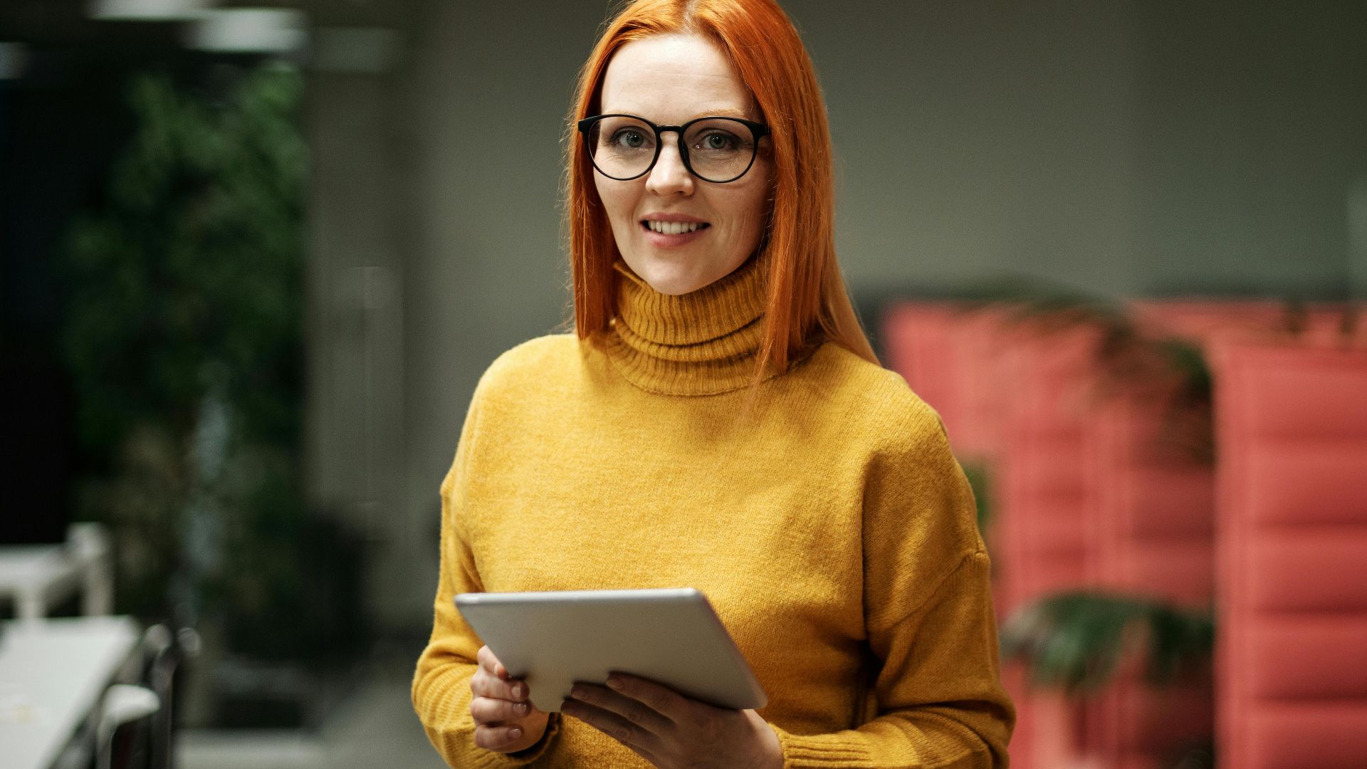 Smiling woman in eyeglasses holding a tablet in a modern office setting.