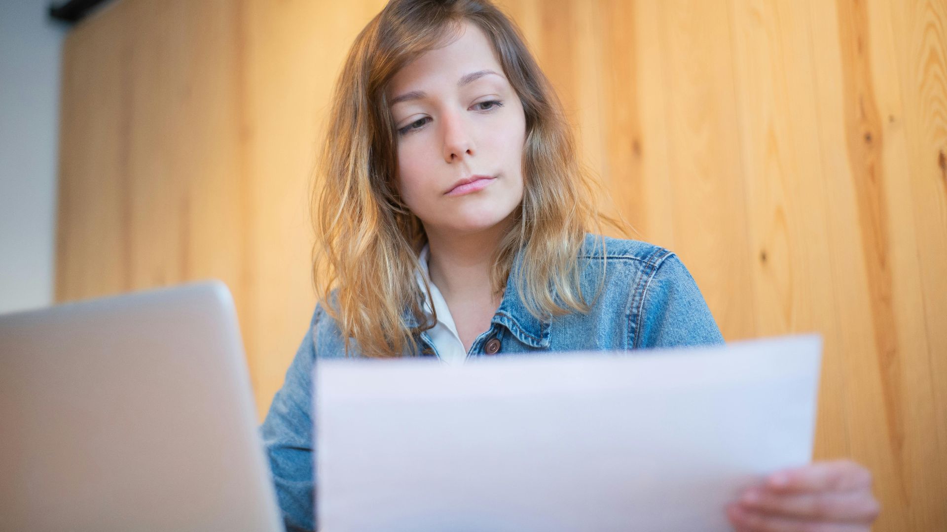 Businesswoman in a denim jacket analyzing documents at a wooden table in a cozy workspace.