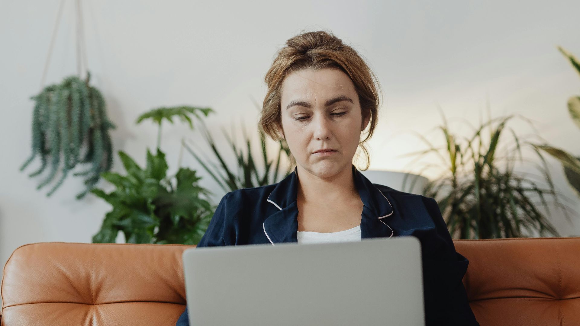 Woman in pajamas working on a laptop in a cozy living room surrounded by plants.