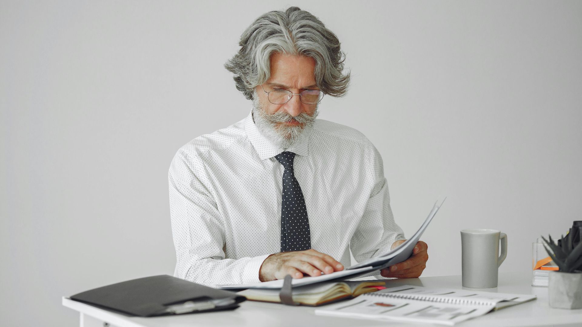An elderly businessman reviewing documents at his desk in a modern office setting.