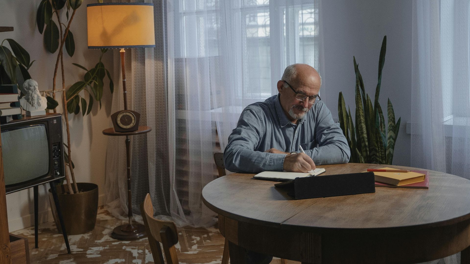 Senior man writing at home in a cozy setting with vintage decor and potted plants.