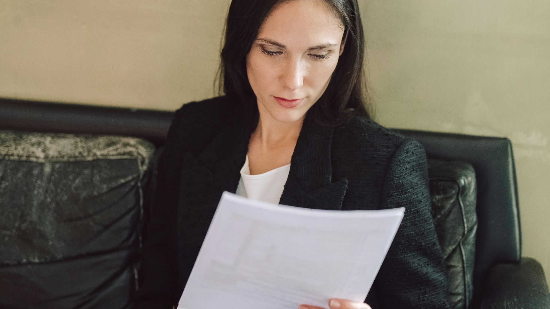 Businesswoman in black blazer analyzing paperwork while sitting on a couch.