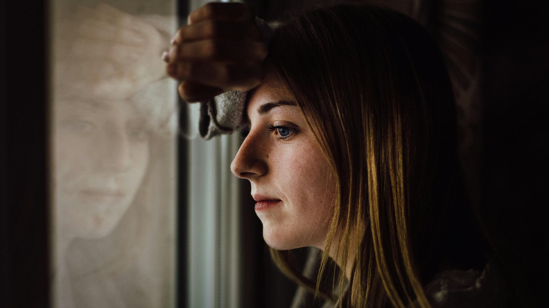 A contemplative young woman looks out a window with a reflective expression, indoors.