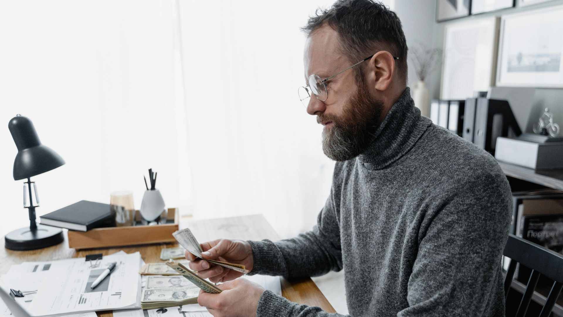 Businessman wearing glasses counting cash at office desk with documents and smartphone.