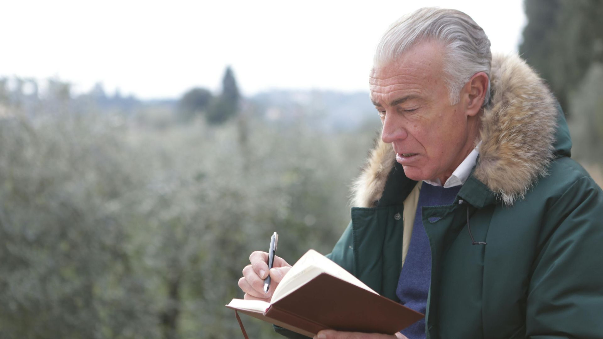 Elderly man writing in a notebook while outside in winter clothing.