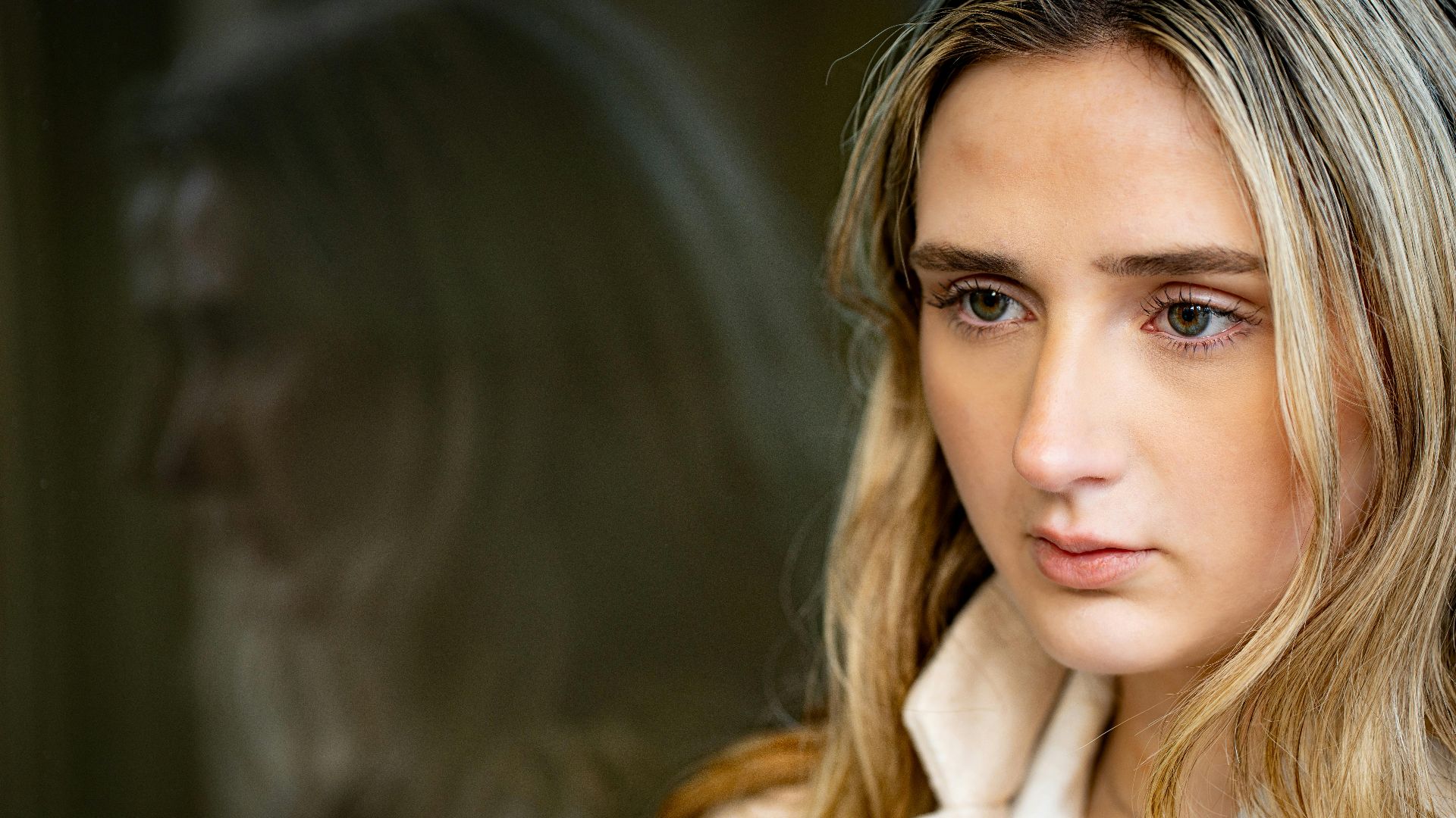 Portrait of a young woman with blonde hair gazing out a window with a reflection.