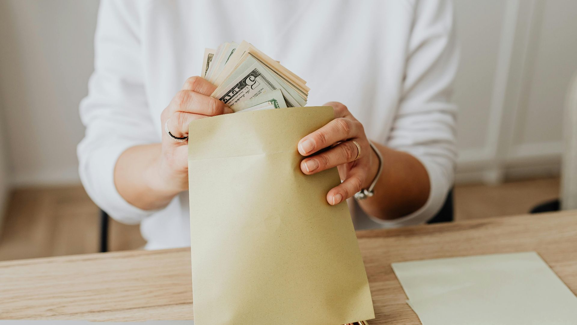 Close-up of person holding envelopes with cash at a wooden desk indoors.
