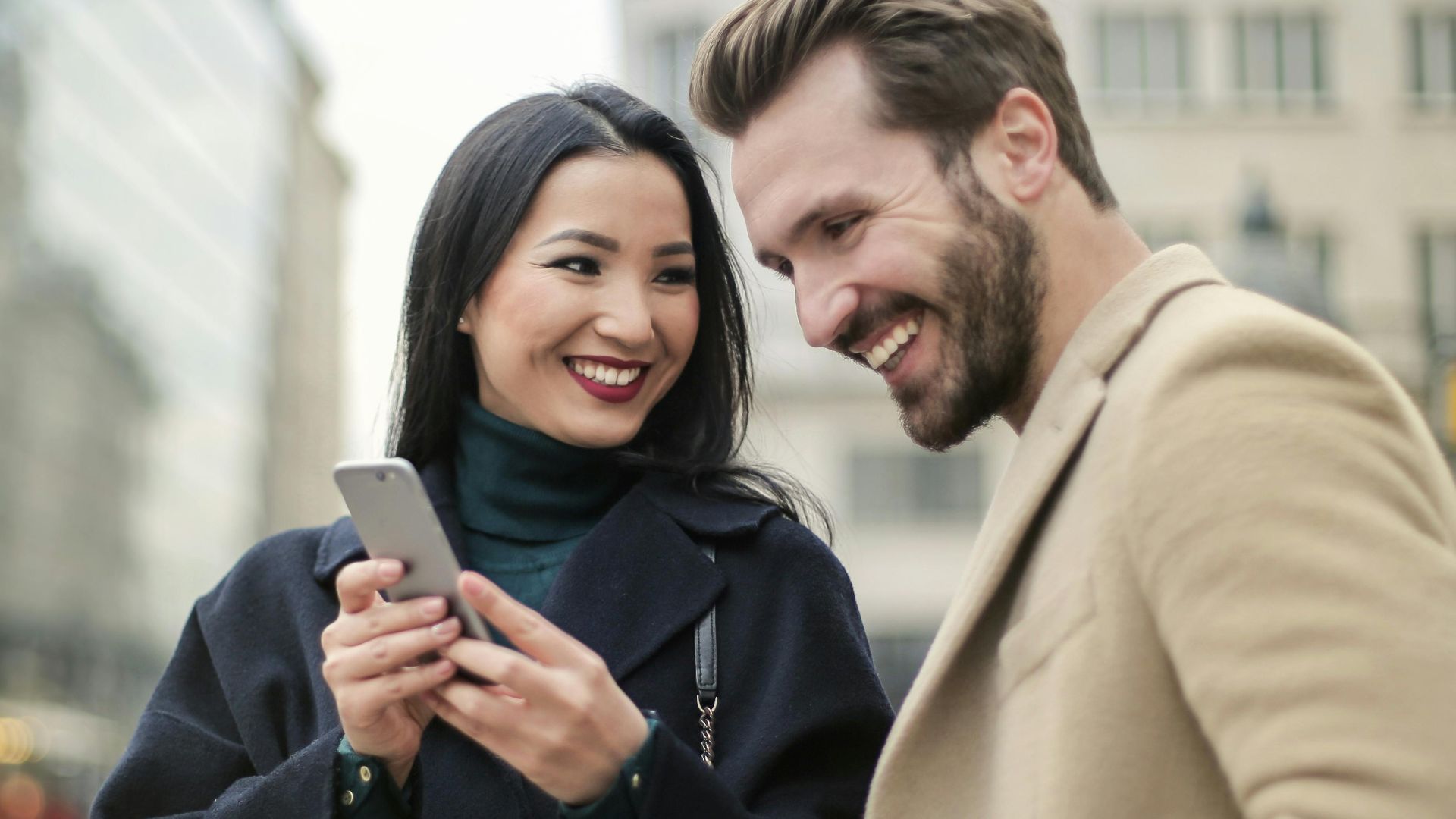 A happy couple shares a joyful moment in the city, interacting with a phone outdoors.