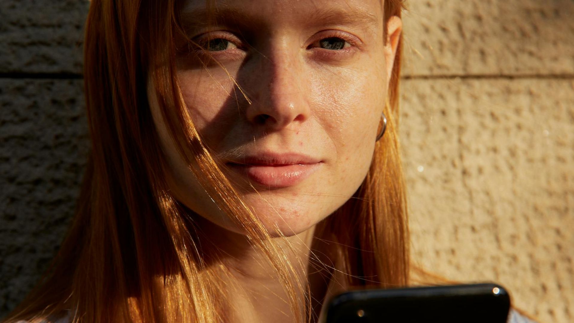 Young woman holding smartphone against a textured wall in warm lighting.