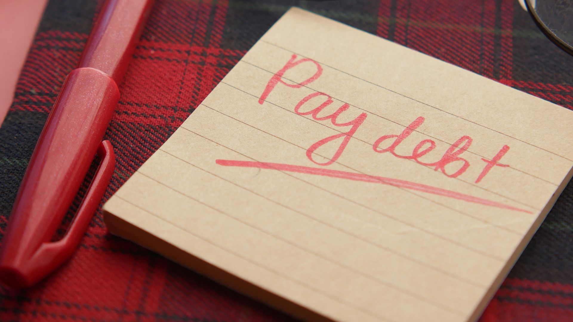 Close-up of a note reading 'Pay debt' next to a red pen on a plaid fabric, emphasizing financial reminders.