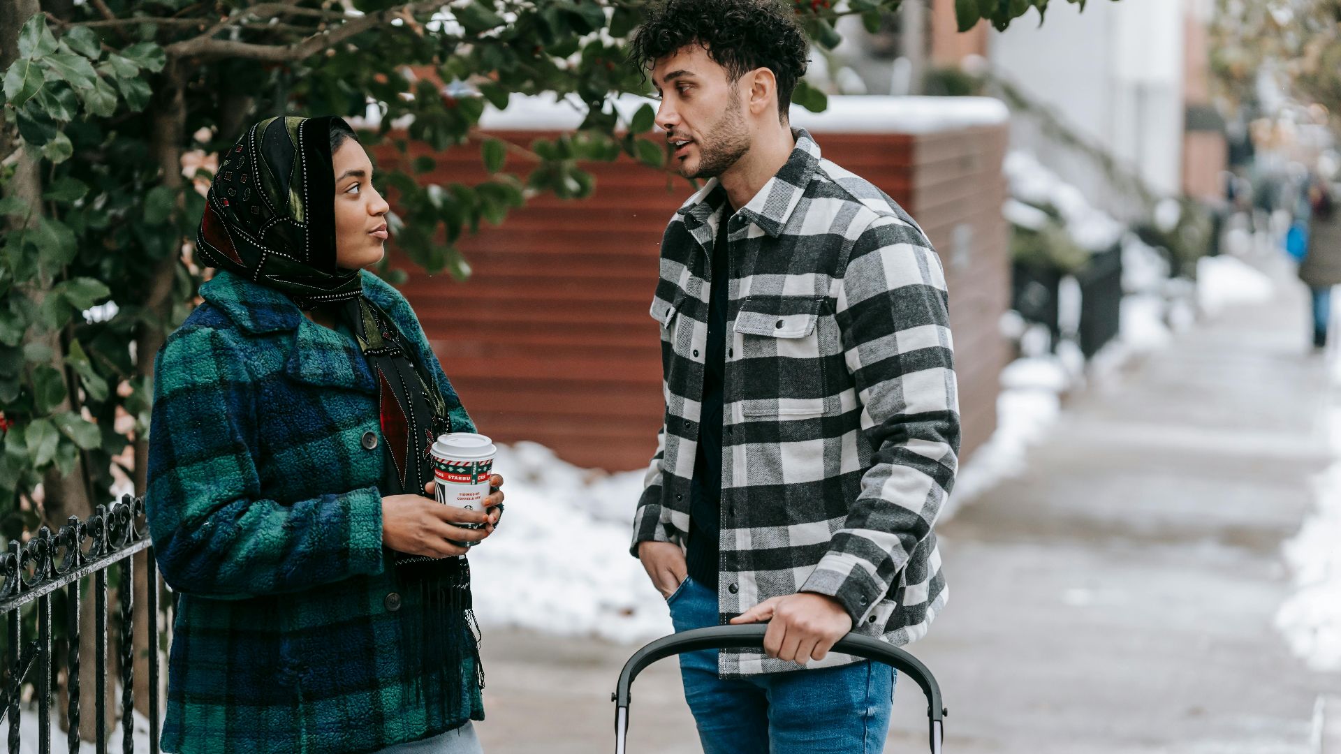 Ethnic woman wearing headscarf standing with cup of takeaway drink and talking to husband standing near stroller on snowy street