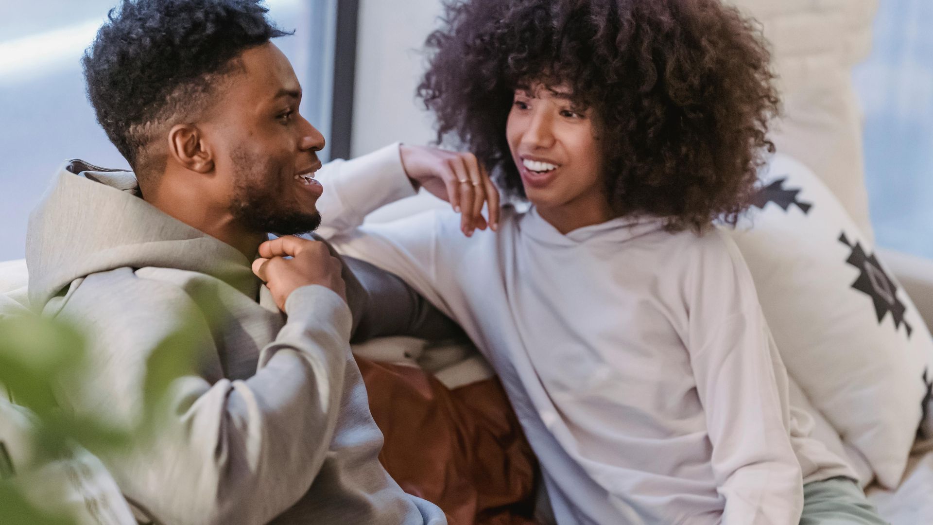 From above of crop positive African American couple smiling and speaking in sofa at home with green plants