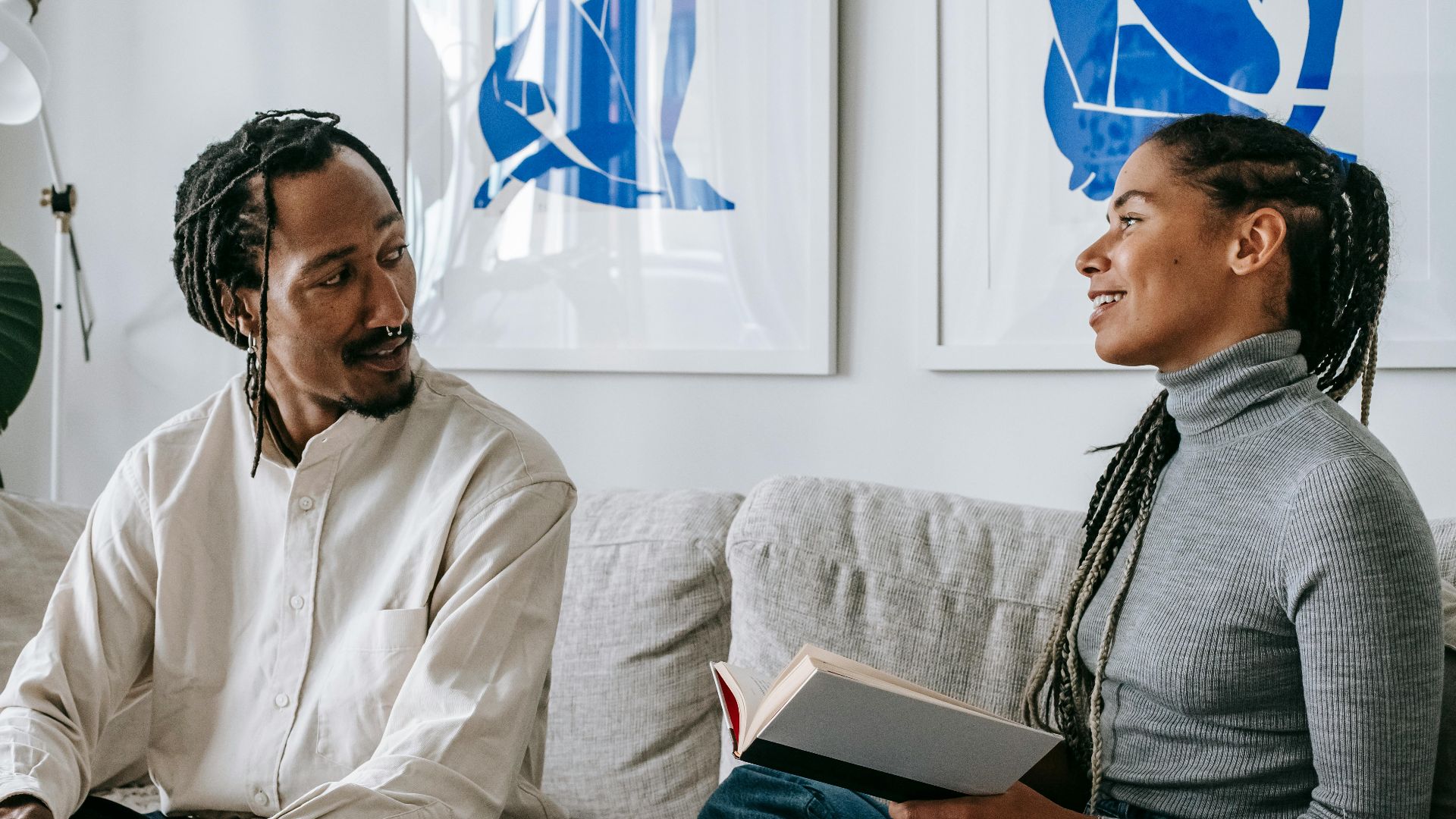 Positive young black lady with Afro braids sitting on sofa with interesting book in hands and speaking with boyfriend working remotely on laptop