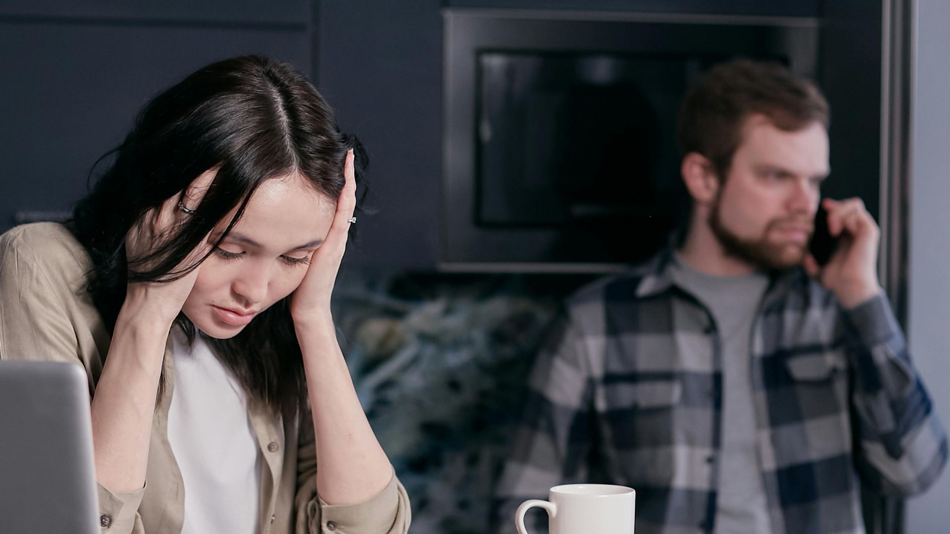 A stressed woman holds her head while working at a desk with documents and a laptop.