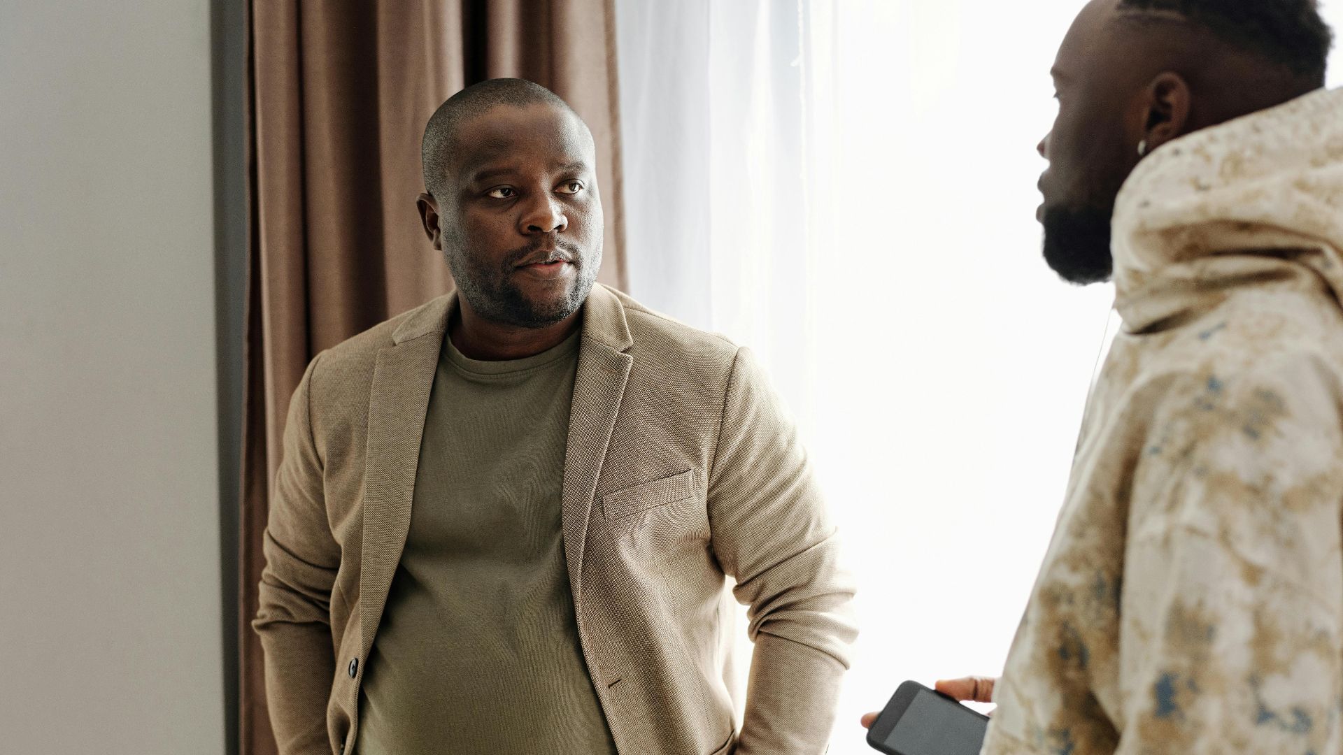 Two men engaged in a serious conversation indoors, one holding a smartphone.
