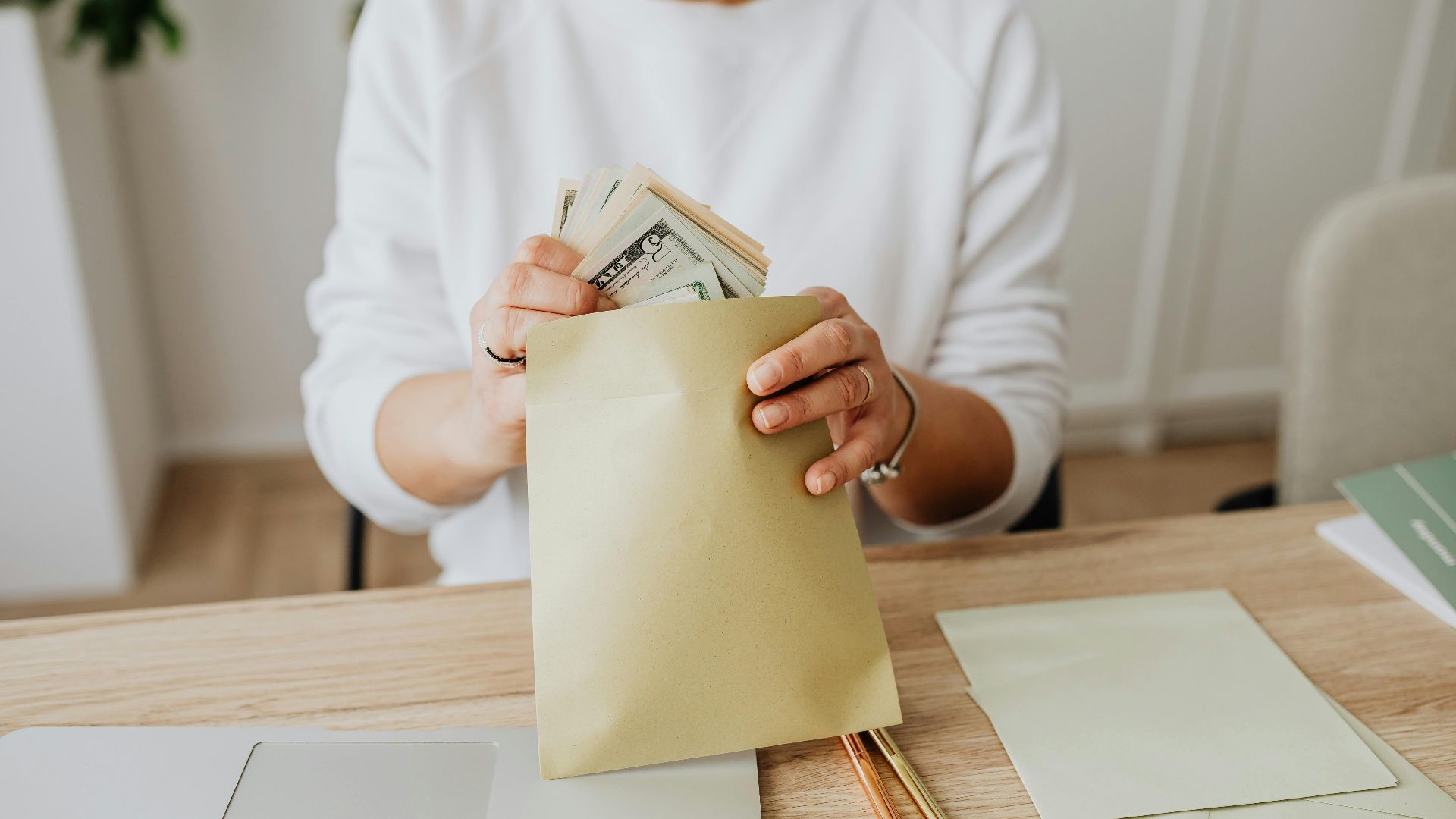Close-up of person holding envelopes with cash at a wooden desk indoors.