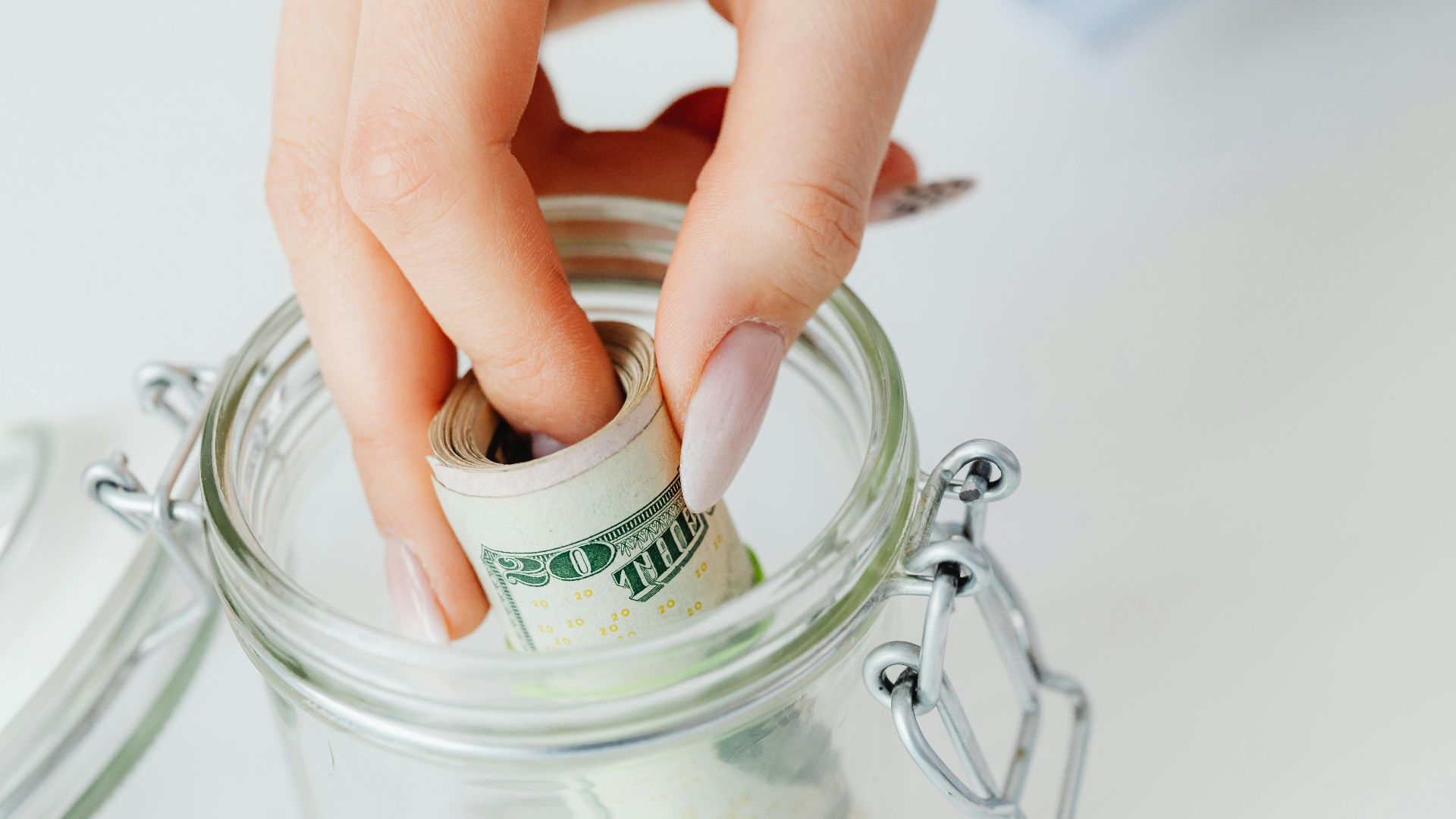 A close-up of a hand placing rolled dollars into a glass jar, symbolizing savings.