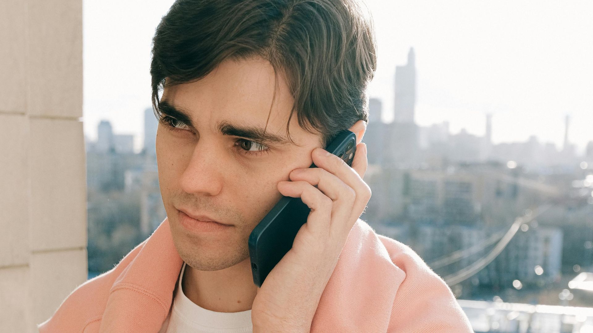 Young man talks on cellphone on an urban balcony with city skyline in the background.