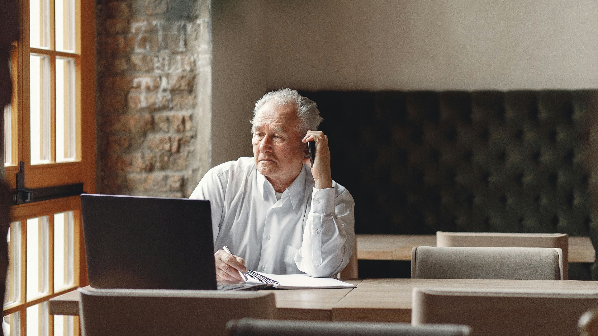 Thoughtfull senior male calling on smartphone and taking notes while working remotely in creative modern workspace at daytime and looking at window