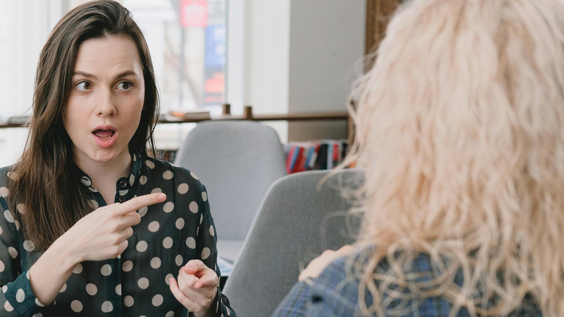 Astonished female pointing away while sitting at table with smartphone and tablet and talking to woman
