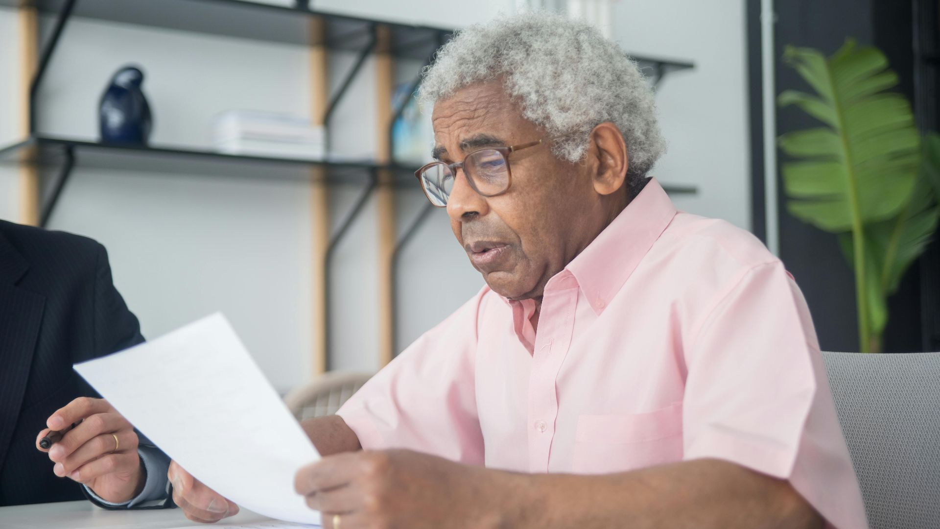 Elderly man with glasses reading a document in a home office setting, reflecting focus and concentration.