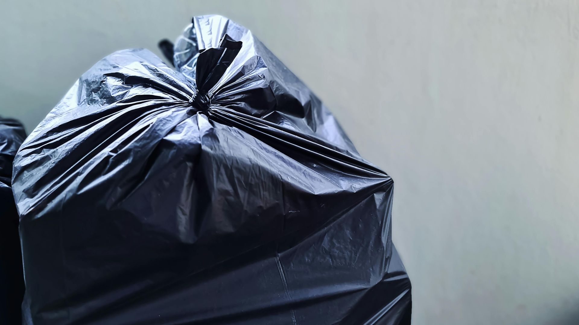 A close-up photograph of a black plastic garbage bag against a plain concrete wall, highlighting waste management.