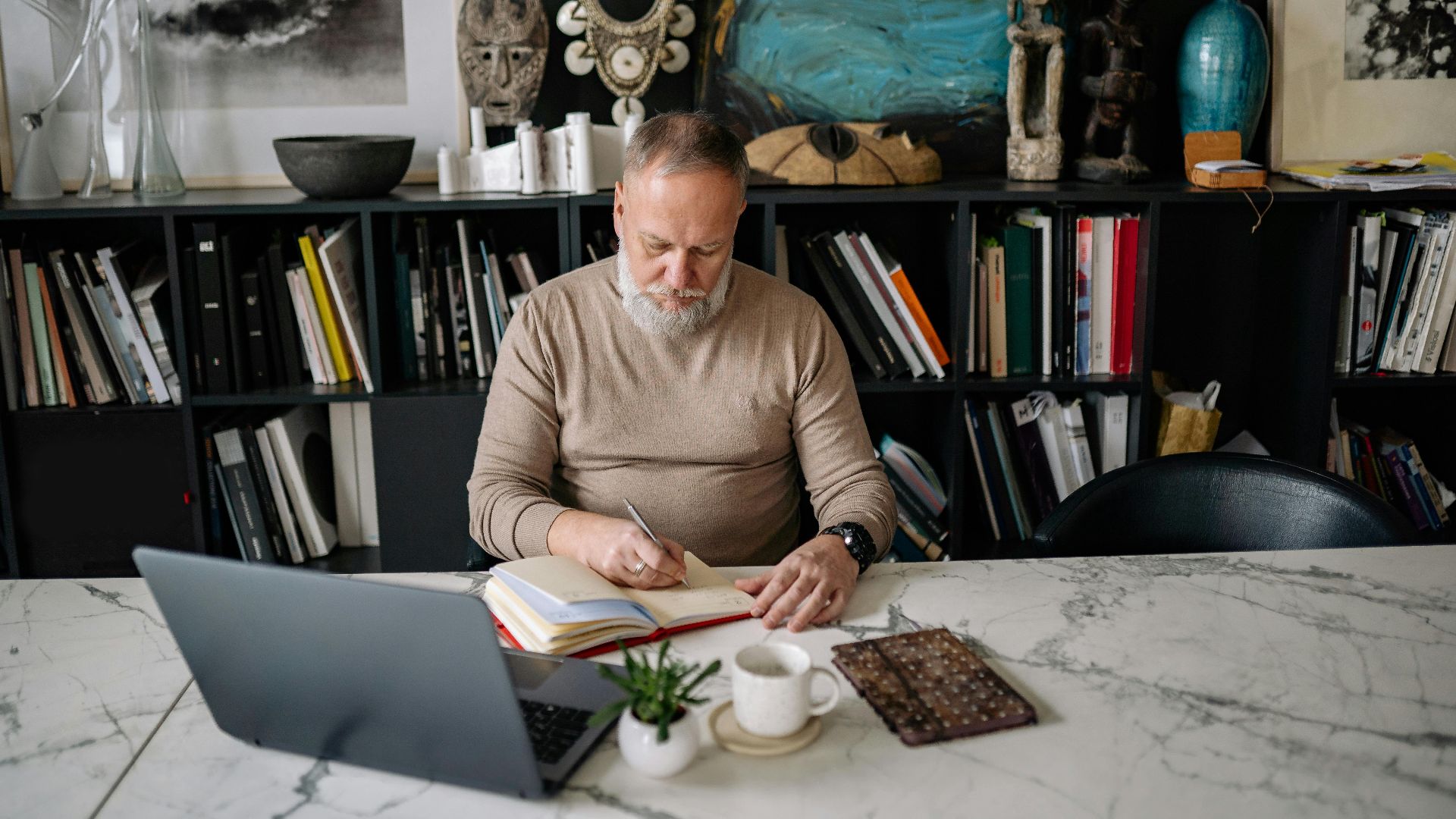 An elderly man with a beard writing in a notebook at a home office desk, surrounded by art and books.