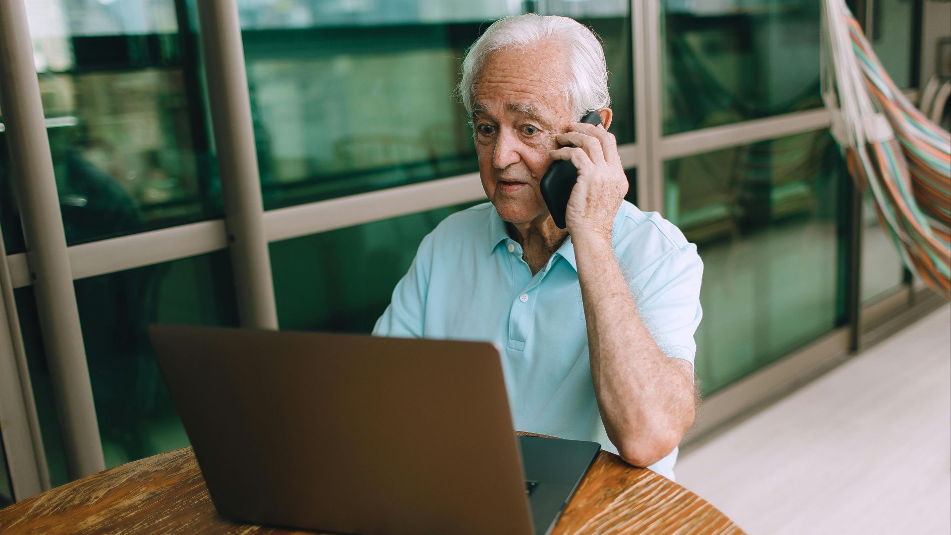Elderly man with gray hair sitting at table using a laptop and talking on the phone indoors.