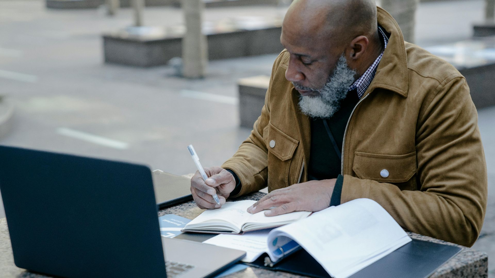 Mature man working with laptop and notebook outdoors, focusing on business tasks.
