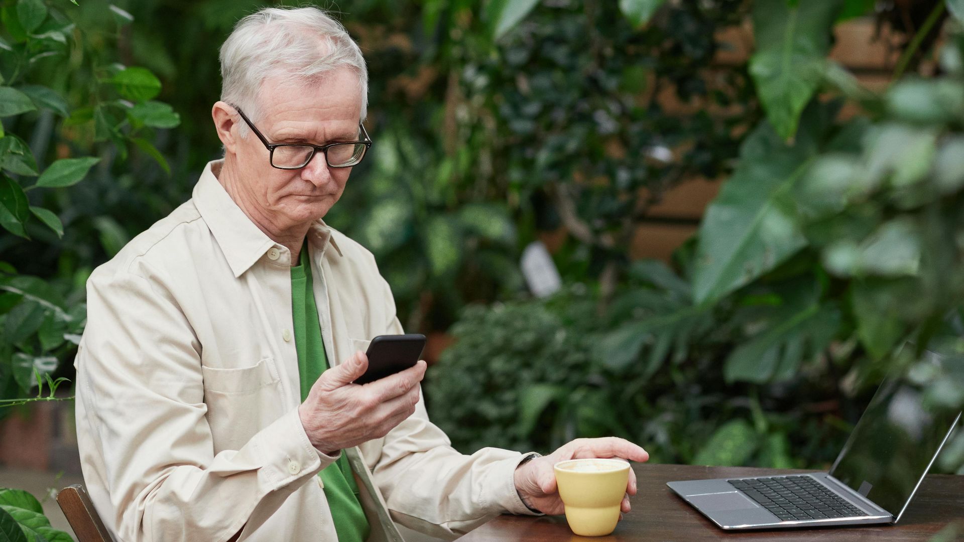 Elderly man using smartphone and laptop, enjoying coffee in a lush garden.