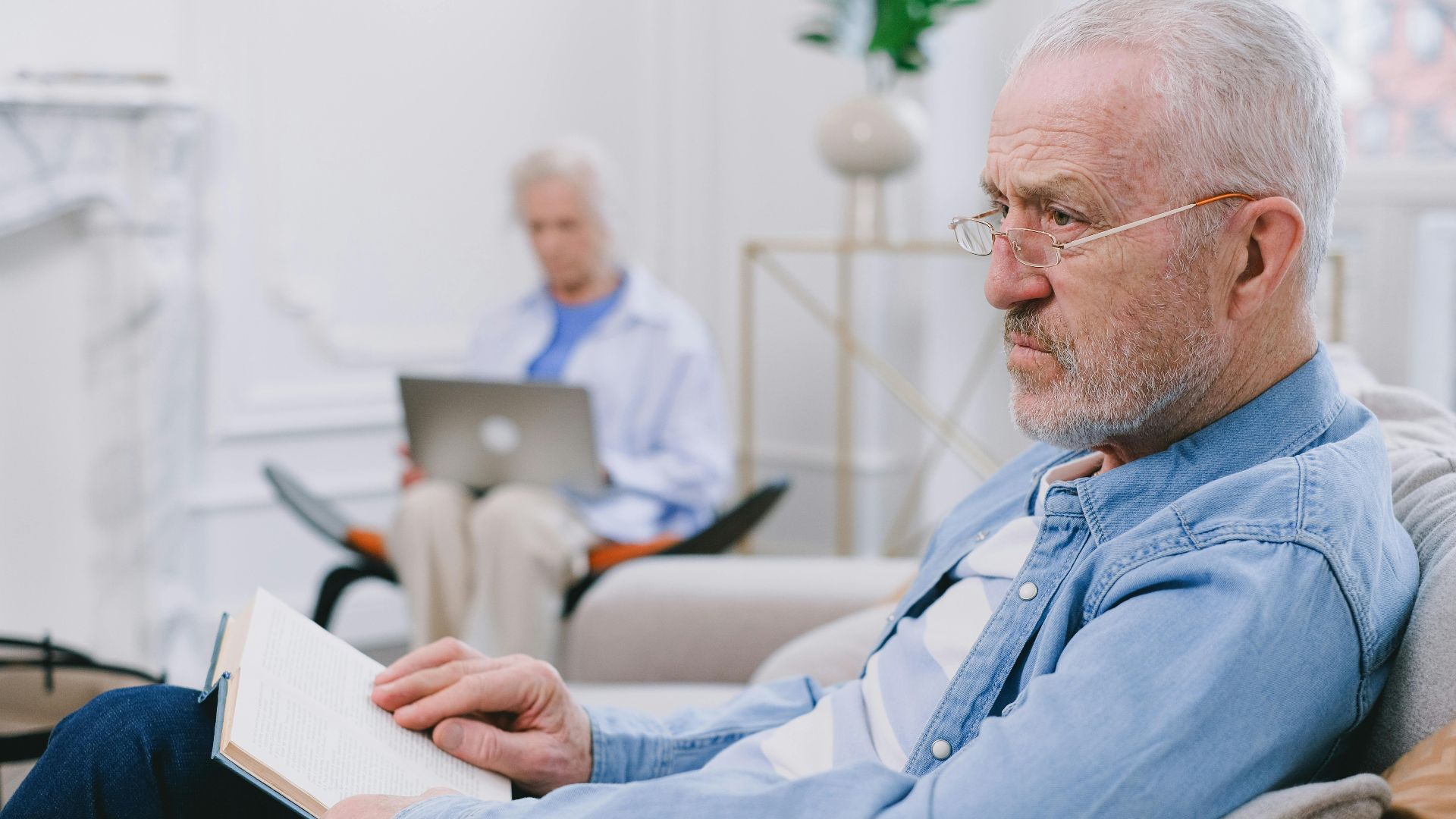 Senior man reading a book while an elderly woman uses a laptop in a cozy indoor setting.